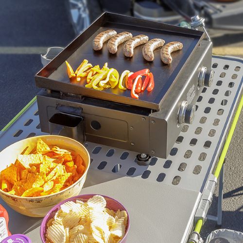 Portable camping griddle on a CORE FlexRail table with sausages and sliced bell peppers cooking, surrounded by bowls of chips on a durable steel cook surface outdoors
