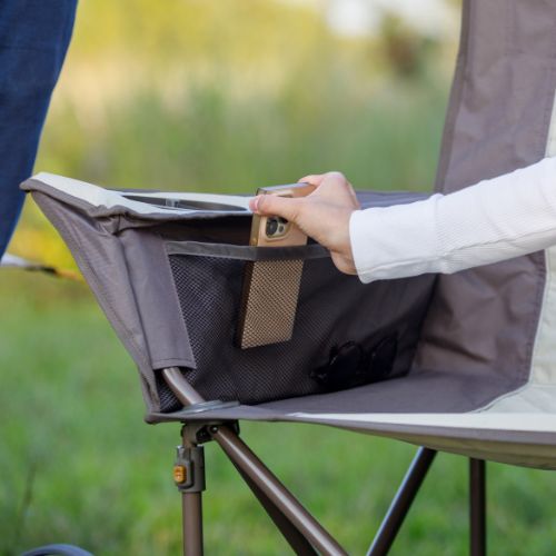 Side armrest of a folding loveseat featuring a built-in storage pocket and cup holder, with a hand placing a smartphone into the pocket; mesh compartments and a sturdy frame are visible against a grassy outdoor background.