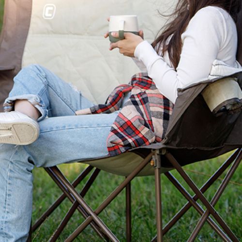 Close-up of a folding loveseat’s metal frame with cross-braced legs with a locking mechanism near the seat, showing sturdy construction and support while set on grass with a blanket draped over the seat.