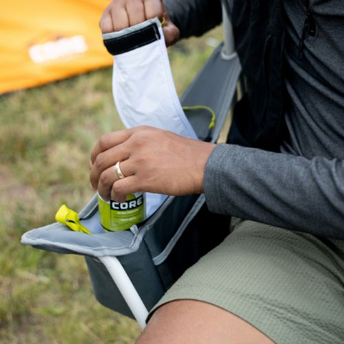 Built-in insulated cooler on the arm of a folding quad chair, with a person placing a tall drink can into the vertical sleeve, set outdoors near a tent.