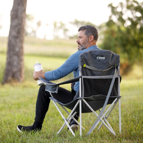 A man sits in a gray folding camp chair on a grassy lawn outdoors, holding a white cup. The chair has a visible CORE logo on the backrest.
