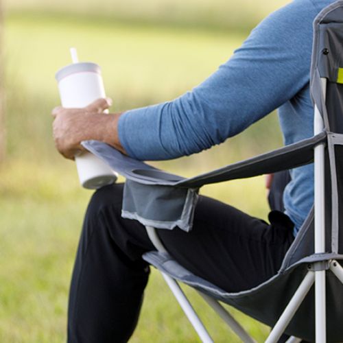 Built-in fabric cup holder on a folding cooler quad chair, with the chair set up outdoors on grass and a seated person’s arm resting nearby holding a drink in a white tumbler.