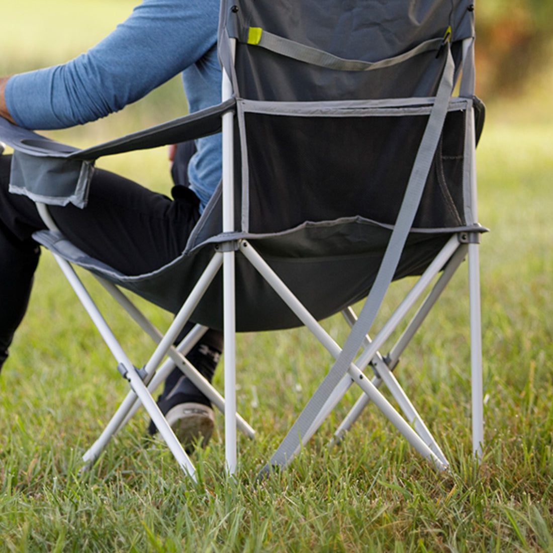 A dark gray folding camp chair on a grassy lawn, with a person sitting in it wearing a blue long-sleeve shirt and black pants.