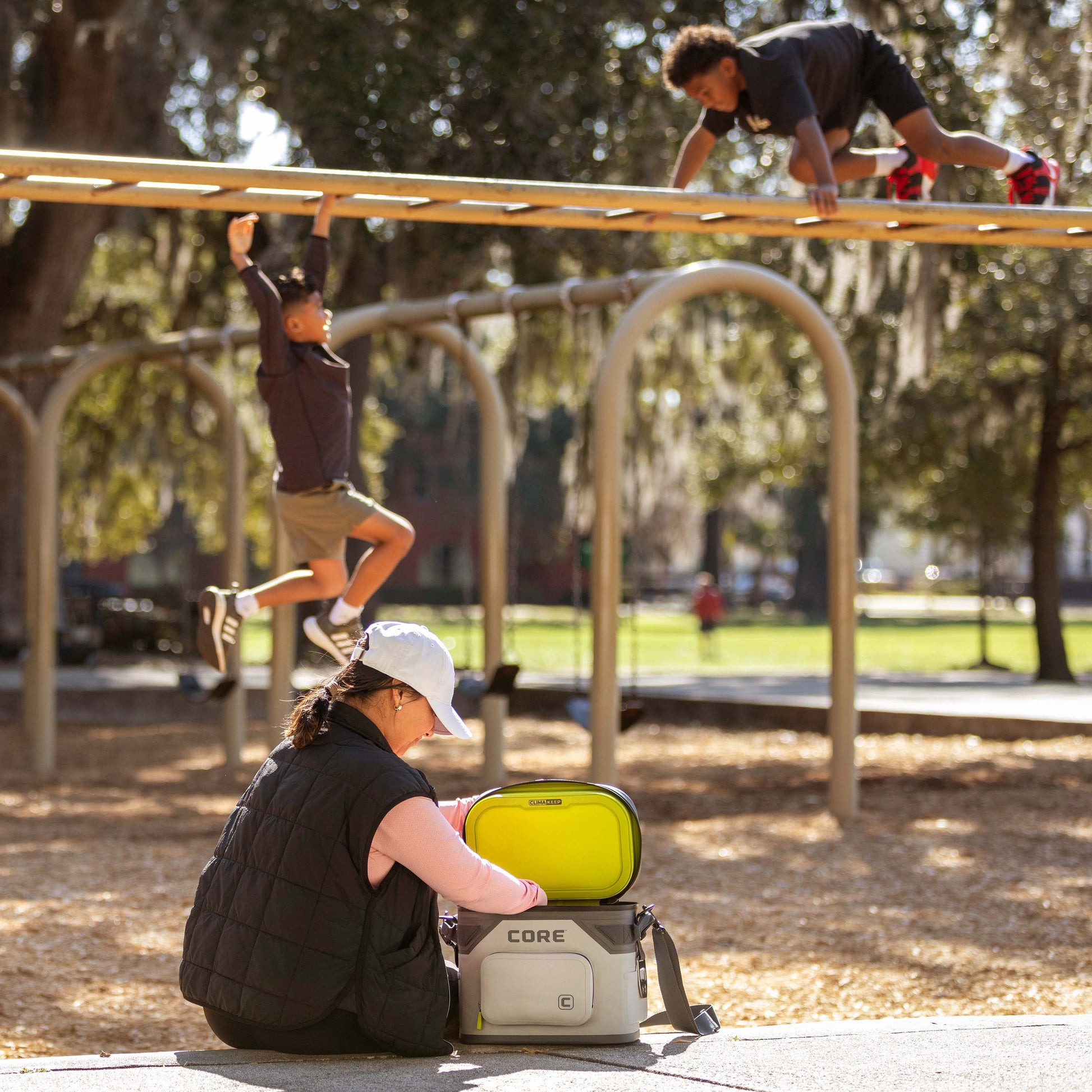 A person with a white cap and black vest is seen opening a gray and yellow Magnetic 12 Can Soft Cooler labeled "CORE" at a park. In the background, two children are climbing and playing on monkey bars under a shaded area with trees.