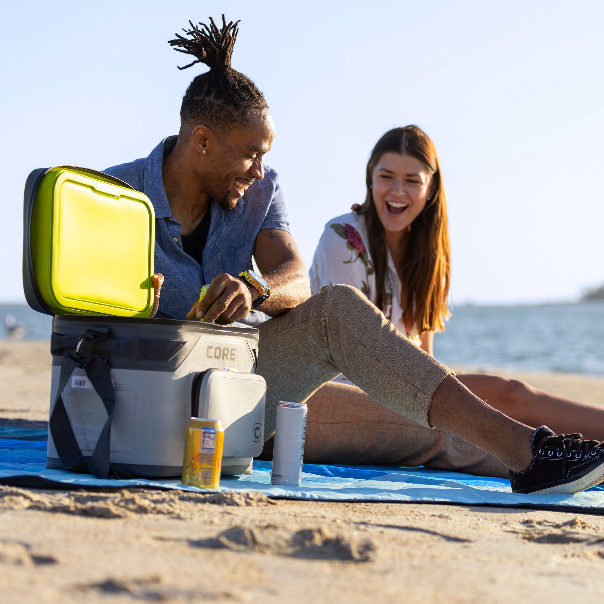 A man and a woman sit on a CORE blue Staydown Outdoor Travel Weighted Ground Blanket at the beach. The man, wearing a blue shirt, opens a CORE Magnetic 18 Can Soft Cooler with a greeen lid, while the woman, with long brown hair, wearing a white floral shirt, laughs. An energy drink and a soda can are placed on the blanket.