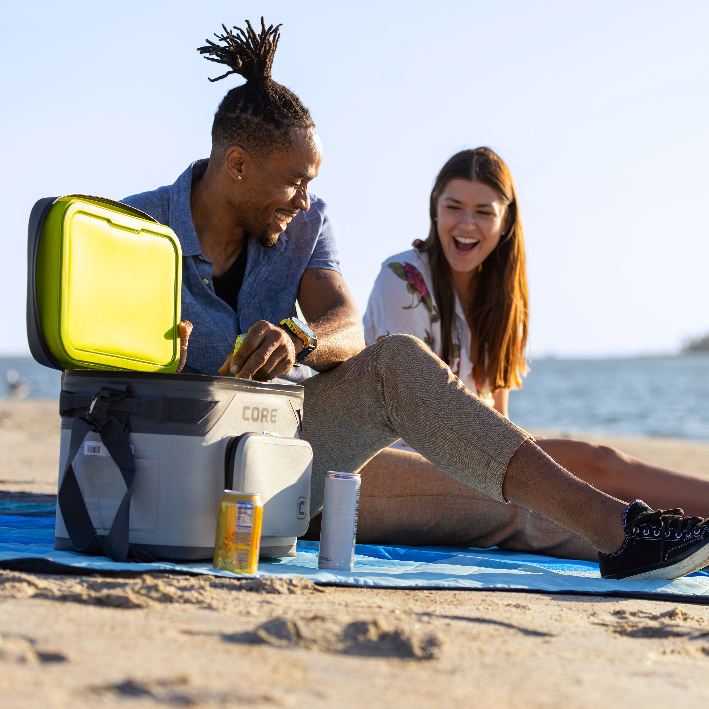 A man and a woman sit on a CORE blue Staydown Outdoor Travel Weighted Ground Blanket at the beach. The man, wearing a blue shirt, opens a CORE Magnetic 18 Can Soft Cooler with a greeen lid, while the woman, with long brown hair, wearing a white floral shirt, laughs. An energy drink and a soda can are placed on the blanket.