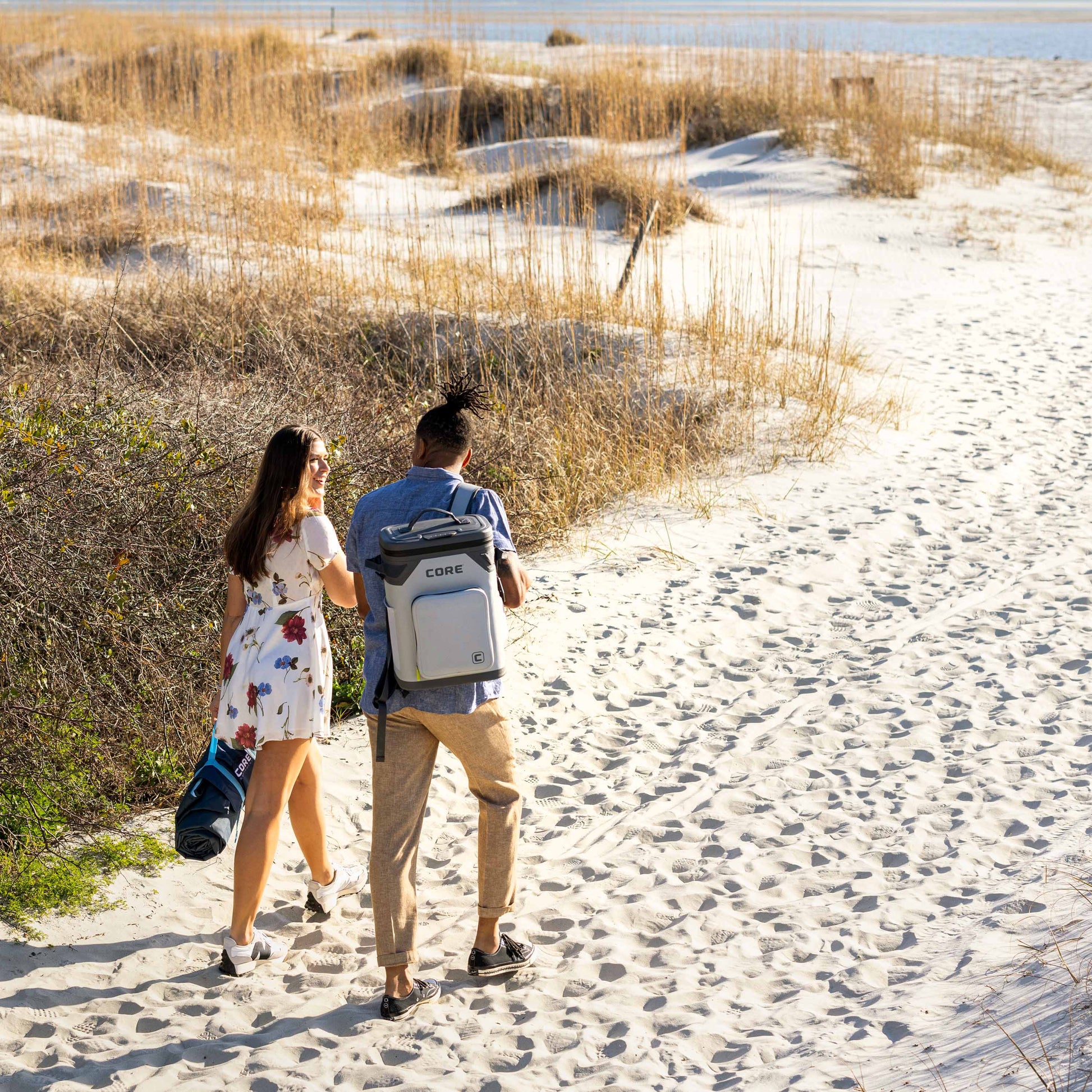 A man and woman walk hand in hand on a sandy beach path towards the ocean. The woman wears a floral dress and carries a CORE Staydown Outdoor Travel Weighted Ground Blanket , while the man wears a blue shirt and beige pants with a grey CORE Magnetic 24 Can Backpack Soft Cooler. The beach is decorated with tall grass and sand dunes.