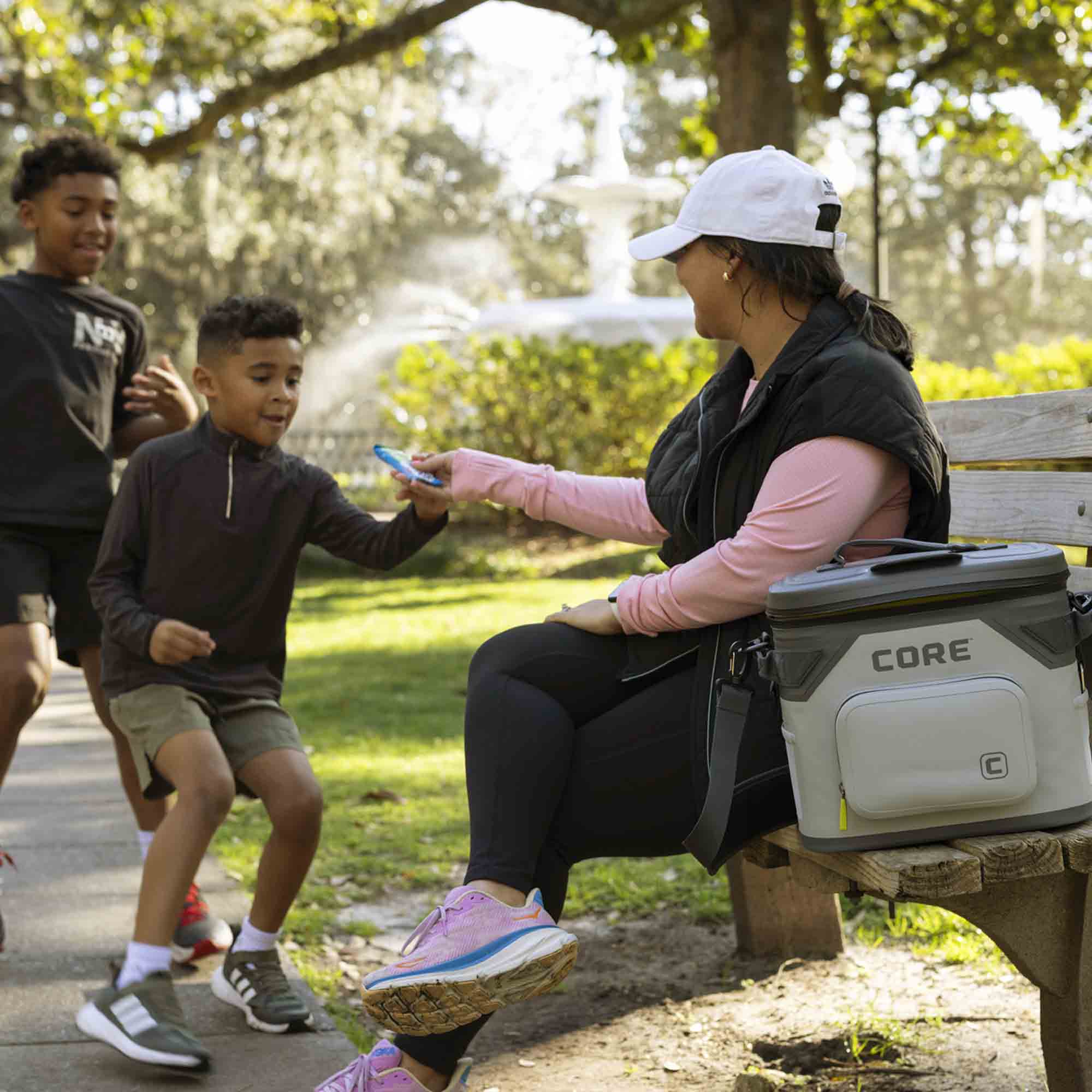 A woman sitting on a park bench with a child, handing them a snack, with a CORE branded Climakeep cooler beside her.