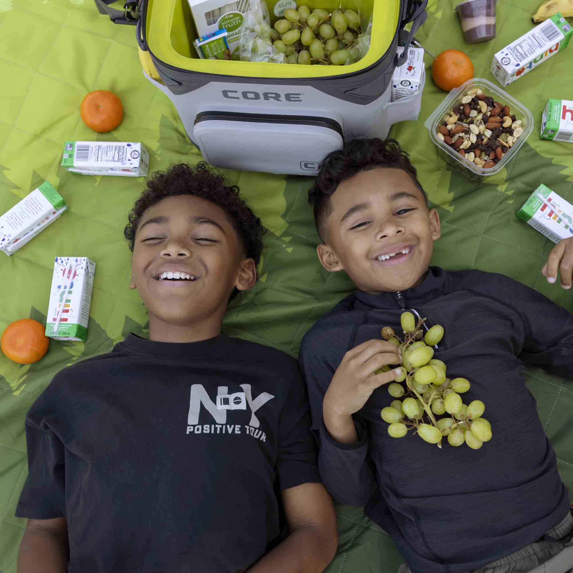 Two children are lying on a green weighted blanket with snacks and a CORE 36 can soft cooler.