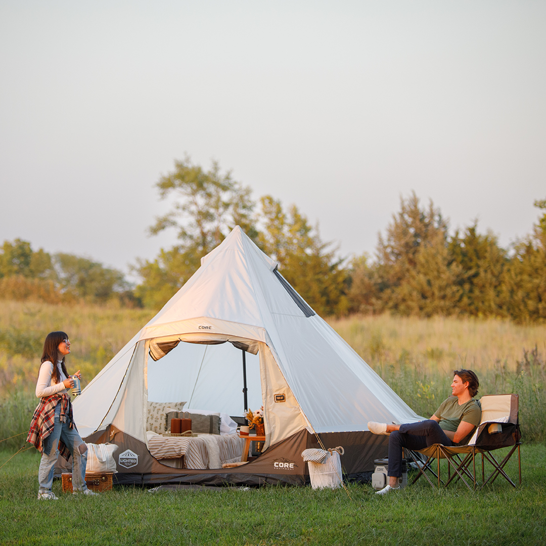 Two people enjoy camping at a peaceful site with a large white CORE 6 person bell tent on the grass, one sitting in a chair, the other standing, surrounded by trees and an open field.