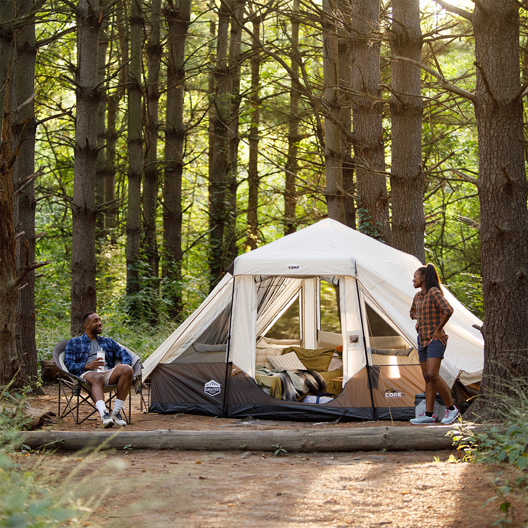 A family enjoys CORE camping gear outdoors, relaxing with a member seated in a chair and another by a spacious CORE 6 person instant pyramid tent amid tall trees, sunlight filtering through for a peaceful, comfortable experience.