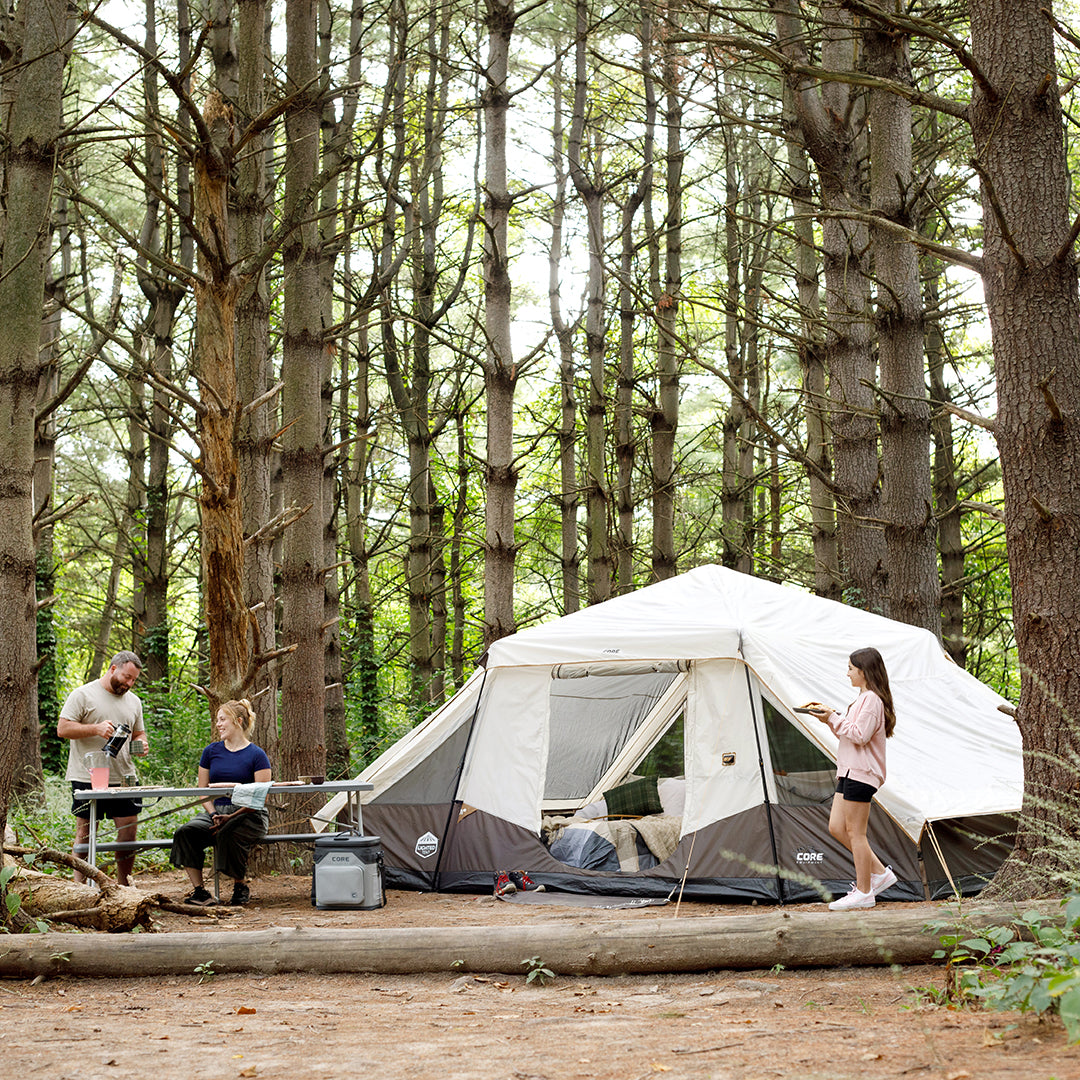 Three people enjoy a camping trip in a forest with a spacious CORE 10 person instant pyramid tent set up among tall pines. One stands by the tent, another sits at a picnic table, and the third prepares food nearby.
