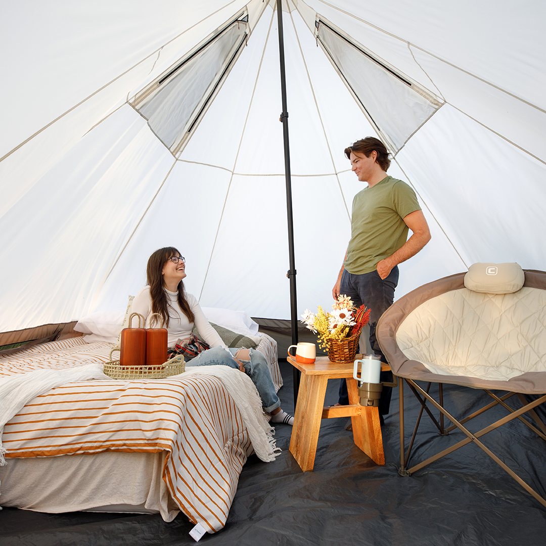 A woman relaxes on a bed while a man stands inside a spacious, well-lit CORE 6 person bell tent. The camping setup includes a bed, cozy chair, and small table with flowers and drinks, providing comfort and an inviting atmosphere.
