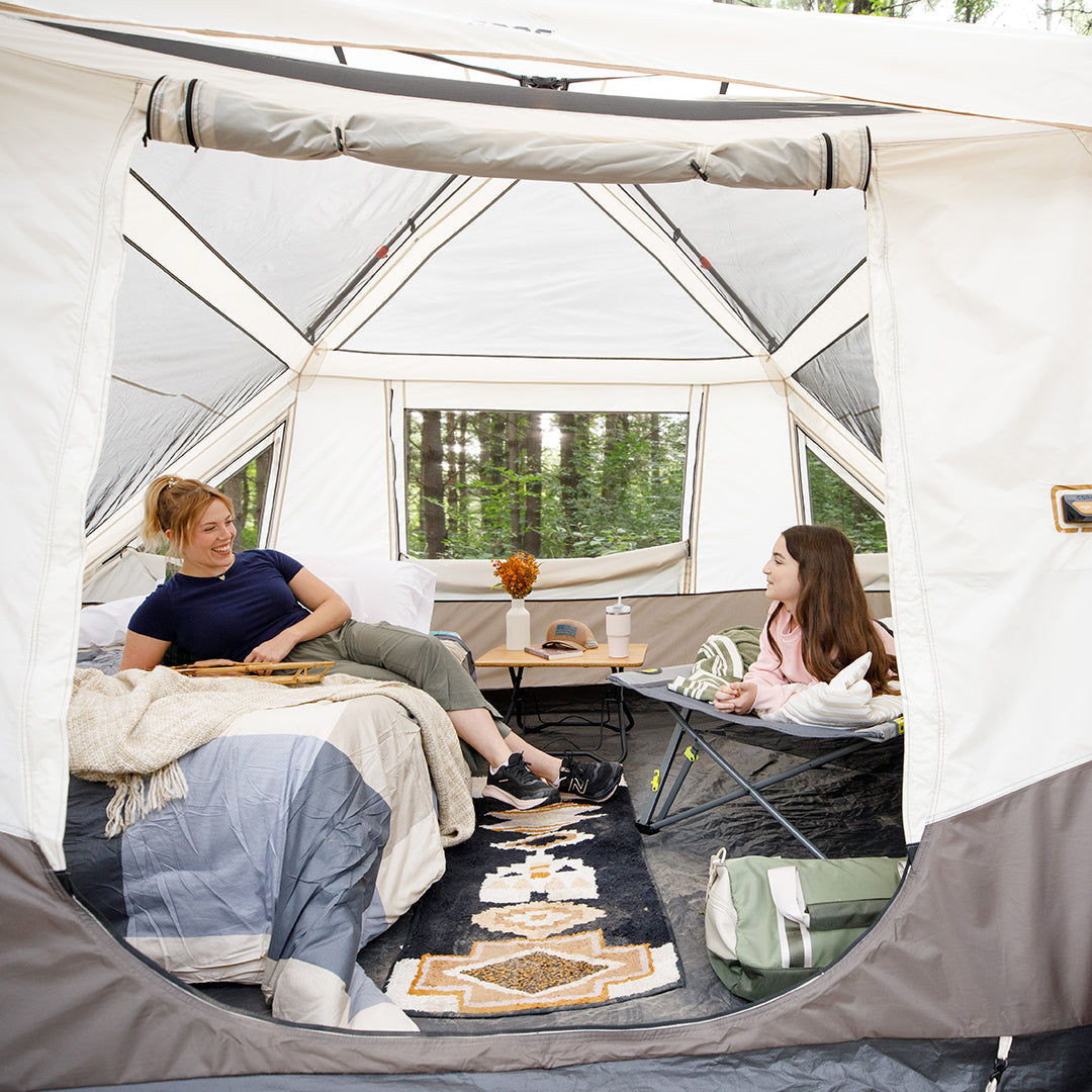 Two people relax in a roomy CORE 10 person instant pyramid  tent with open windows. One sits on a bed, the other on a camp chair by the table. Cozy bedding and outdoor gear surround them as they chat, with trees visible outside.