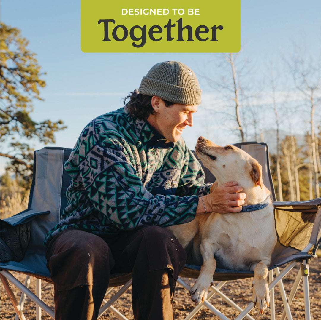 A person wearing a patterned jacket and a knit beanie is sitting on an outdoor chair, lovingly holding a dog on their lap. The sky is clear, trees are visible in the background. Above, text reads "Designed to be Together." They're sitting on the CORE Loveseat Double Outdoor Camp Chair.