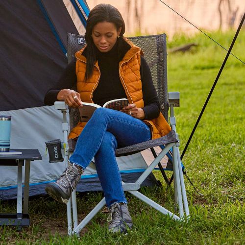 A woman sits in a CORE Stretch Knit Hard Arm Chair outdoors next to a tent, reading a book. She is wearing an orange quilted vest over a black top, blue jeans, and gray boots.