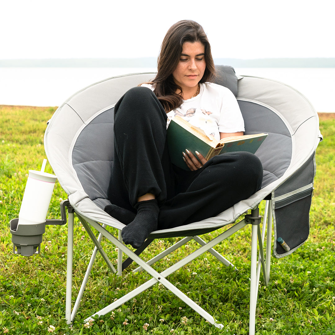 A woman sits in a gray and white folding camp chair on a grassy lawn by the water, reading a green book. The chair has a side cup holder with a white cup.