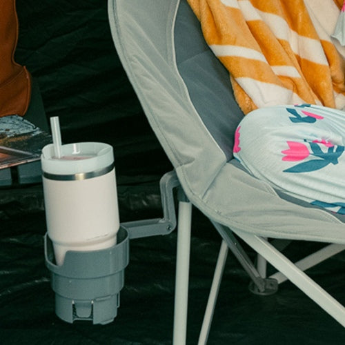 Portable cup holder clipped to the arm of a folding outdoor chair, holding a large white tumbler with a straw, set on green grass.