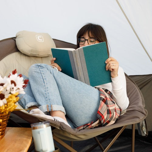 A woman wearing glasses relaxes in a camping chair inside a tent, reading a teal book. She is dressed in light blue jeans and a white long-sleeve top, with a plaid blanket draped over her lap.