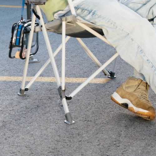 Folding outdoor chair with wide, all-terrain feet providing stability on pavement, shown with crossed metal legs and a seated person’s legs and shoes visible nearby.