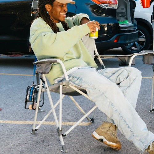 A person sitting in a folding camp chair in a parking lot, wearing a light green sweatshirt, light blue jeans, and tan boots, holding a drink.