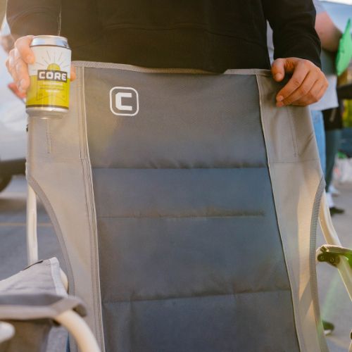 Close-up of a CORE XL Padded Tension Chair featuring a plush, quilted seat and backrest, with visible padded stitching, a built-in cup holder holding a canned drink, and a person’s hands resting on the chair in warm outdoor lighting.