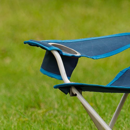 Built-in mesh cup holder on the arm of a blue folding youth chair, attached to a padded armrest and positioned above crossed metal frame legs on green grass.