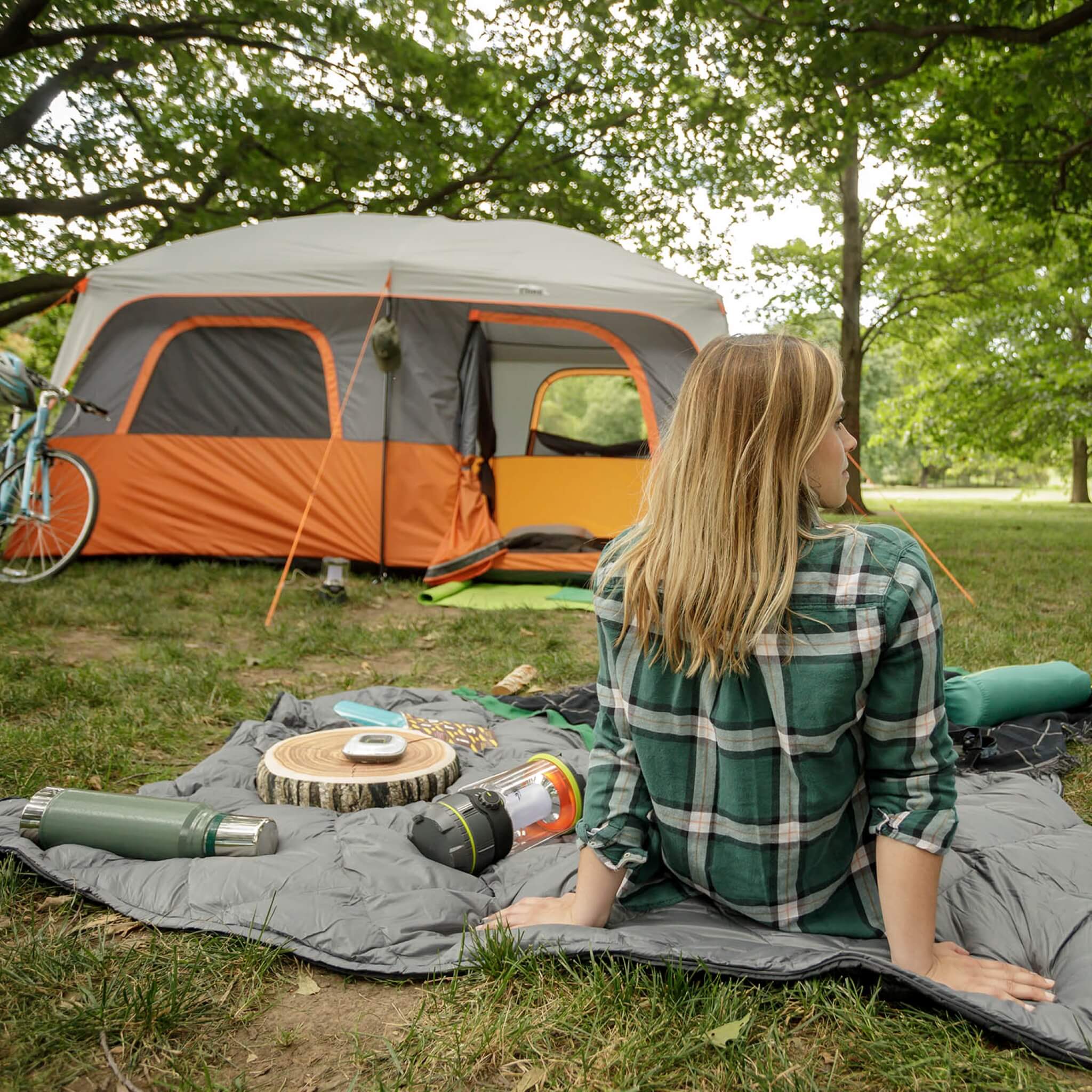 CORE 10 person straight wall cabin tent in orange and gray set up at a campsite with camper relaxing under trees beside the tent.