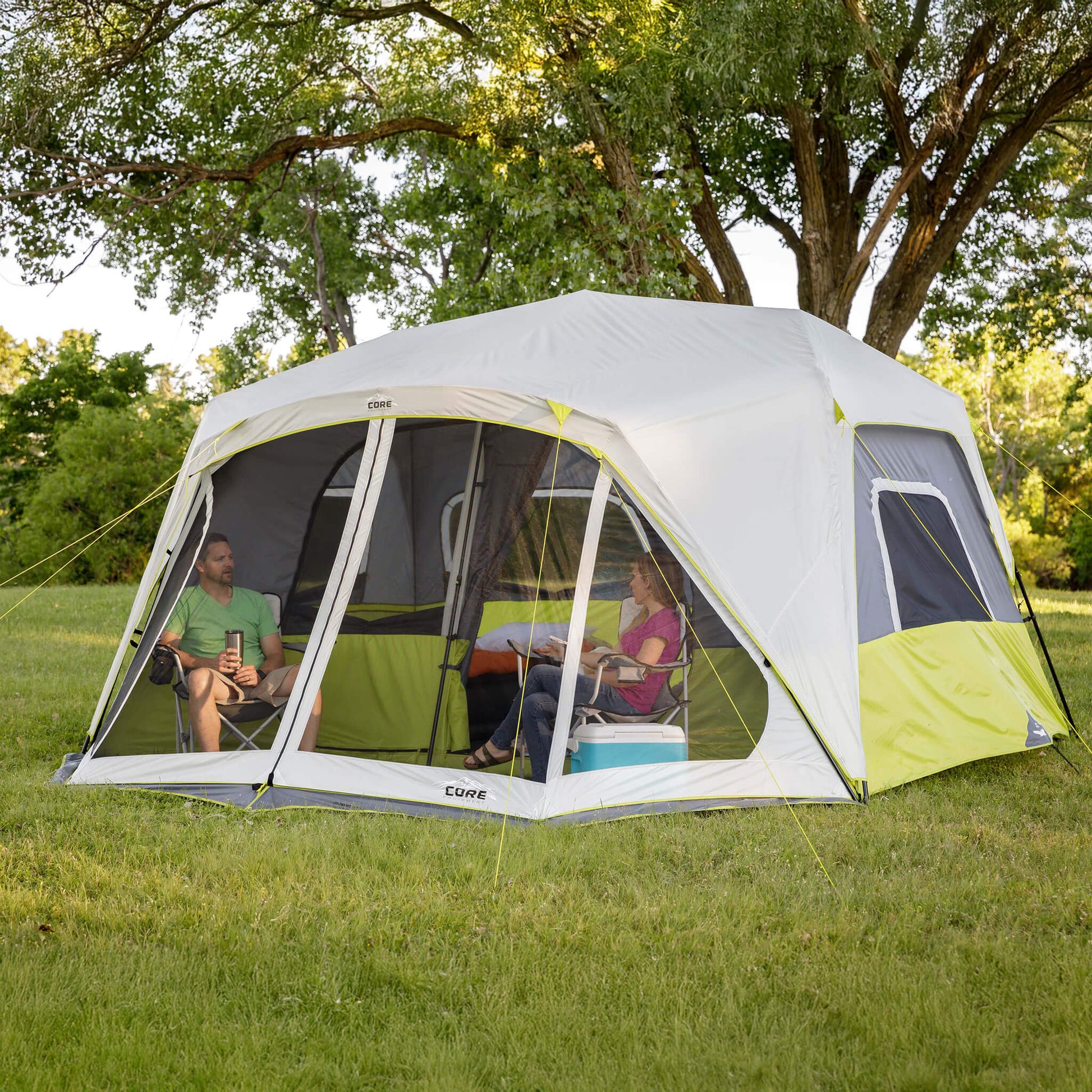 Campers relaxing beside the CORE 10 person instant cabin tent with screen room in gray and green, designed for fast setup and reliable weather protection.