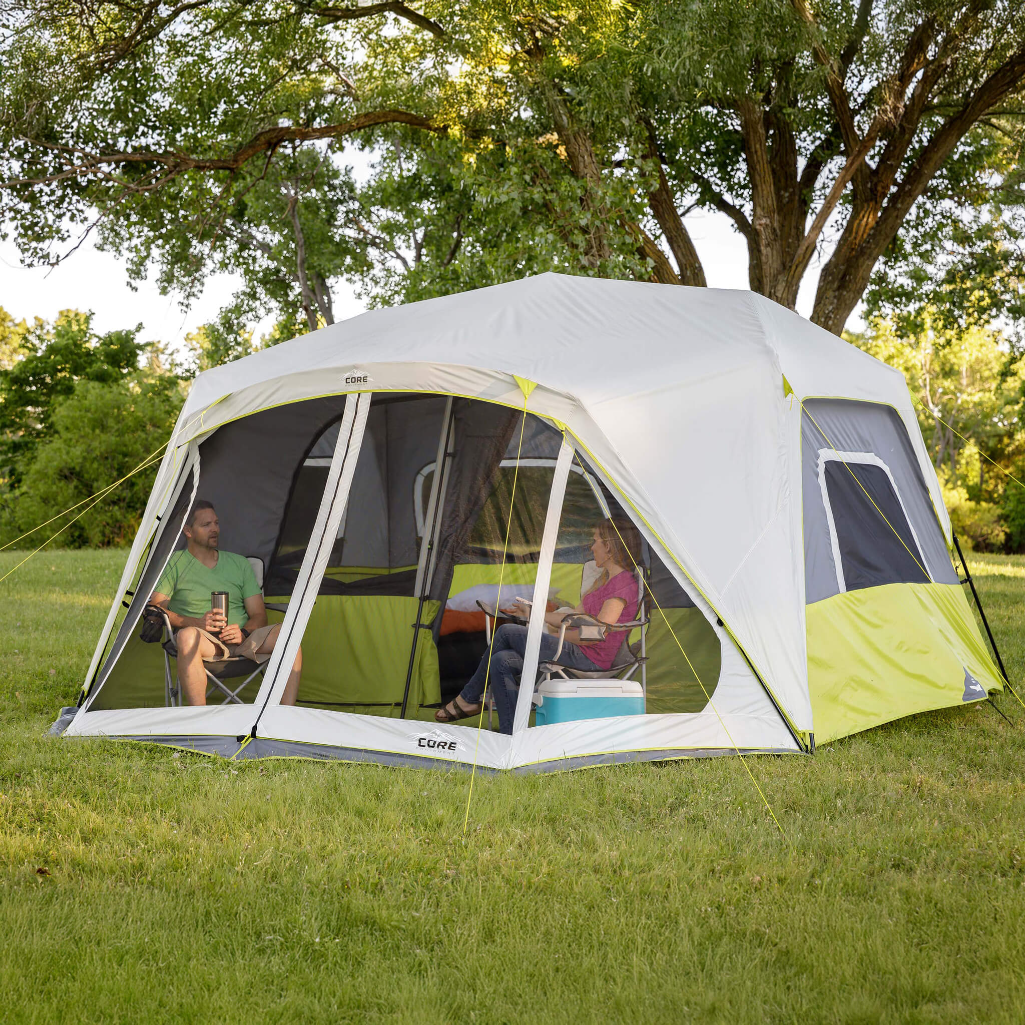 Campers relaxing beside the CORE 10 person instant cabin tent with screen room in gray and green, designed for fast setup and reliable weather protection.
