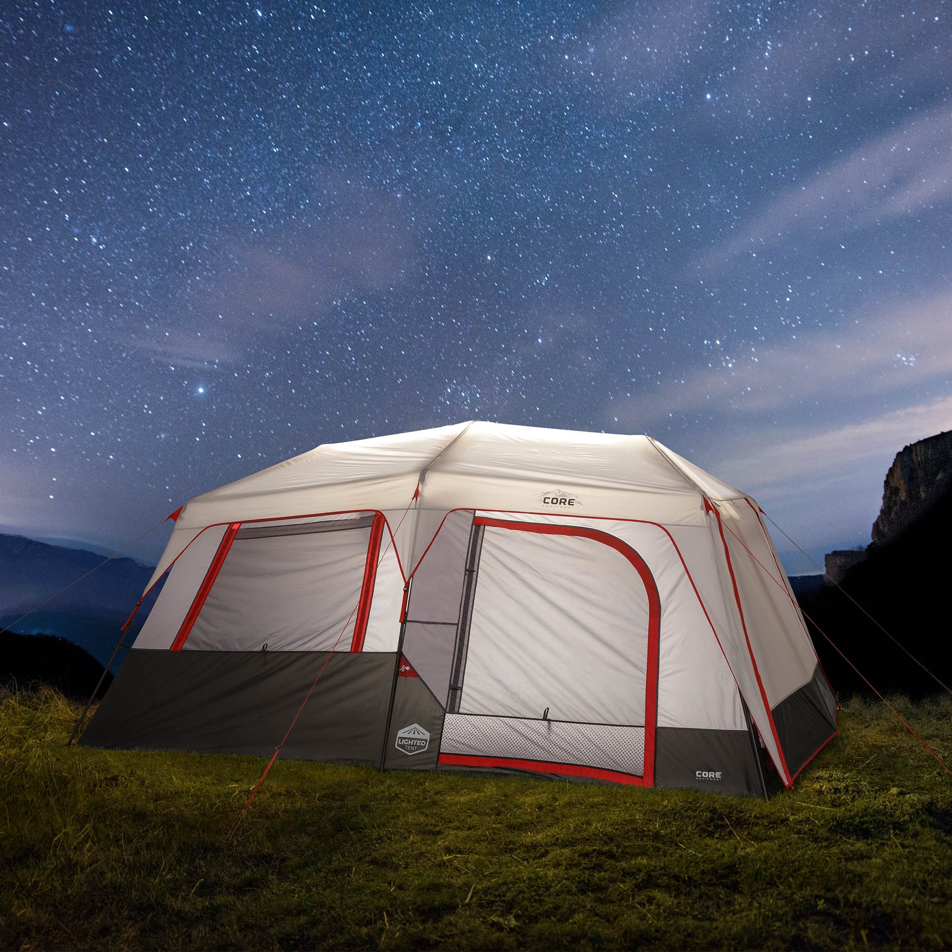 A large white and gray CORE camping tent with red trim pitched on grass under a star-filled night sky.