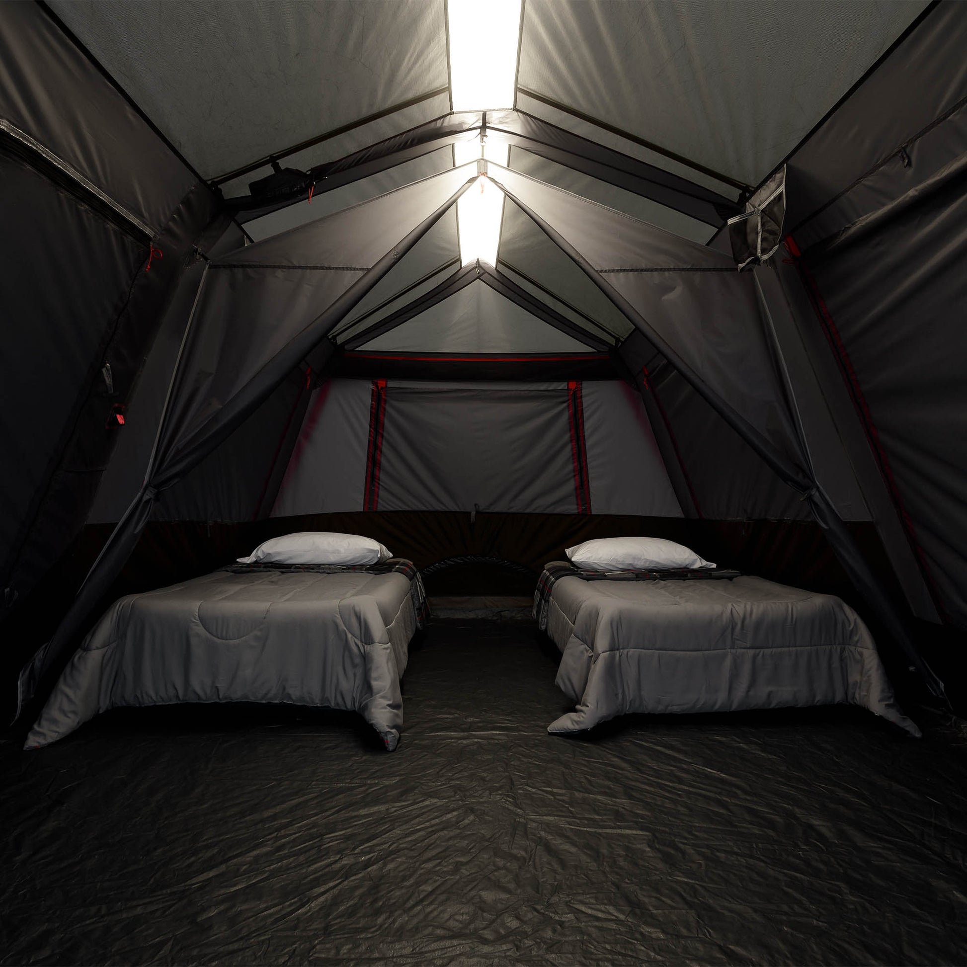 The interior of a large camping tent with two single beds, gray bedding, and white pillows, illuminated by bright rectangular lights at the peak.