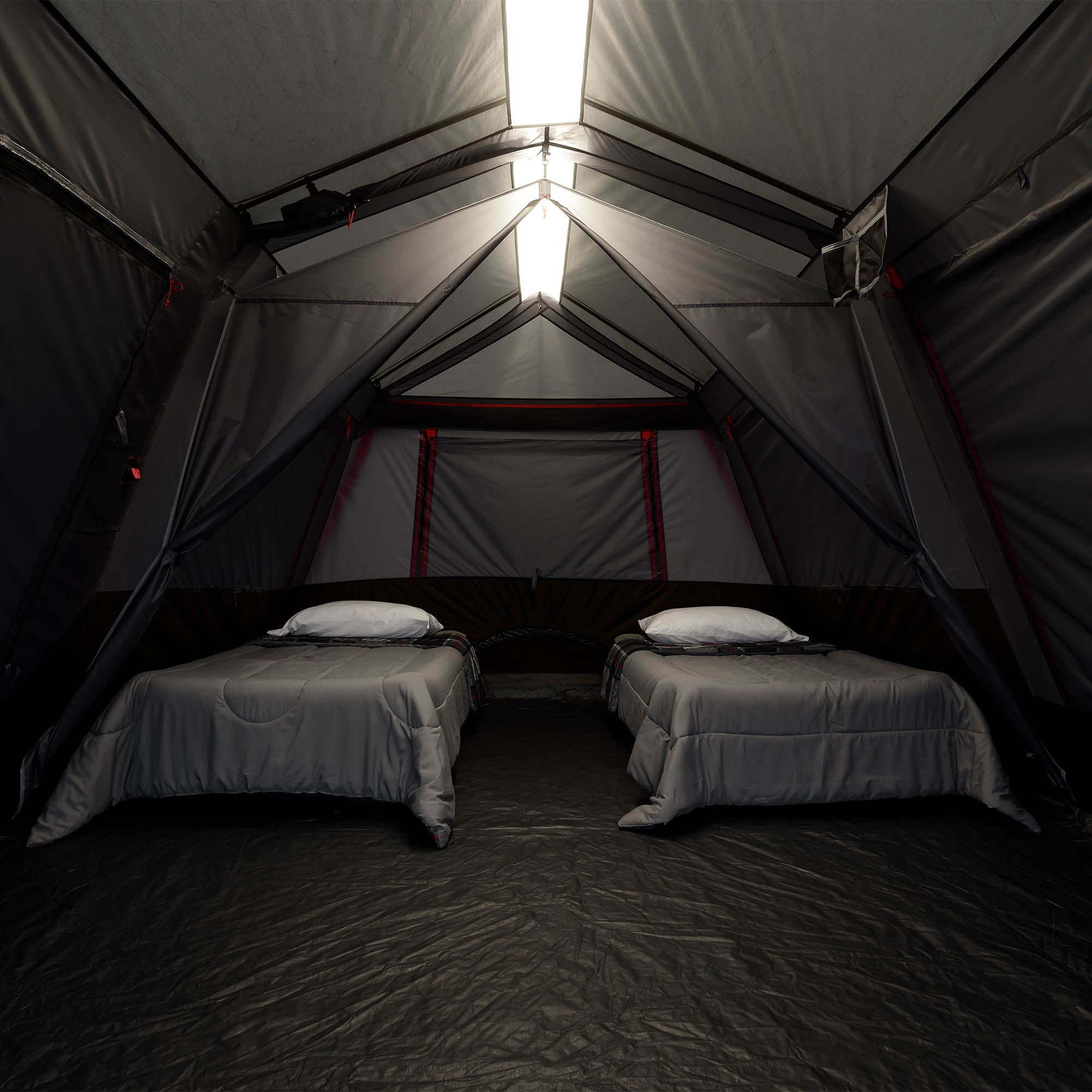 The interior of a large camping tent with two single beds, gray bedding, and white pillows, illuminated by bright rectangular lights at the peak.