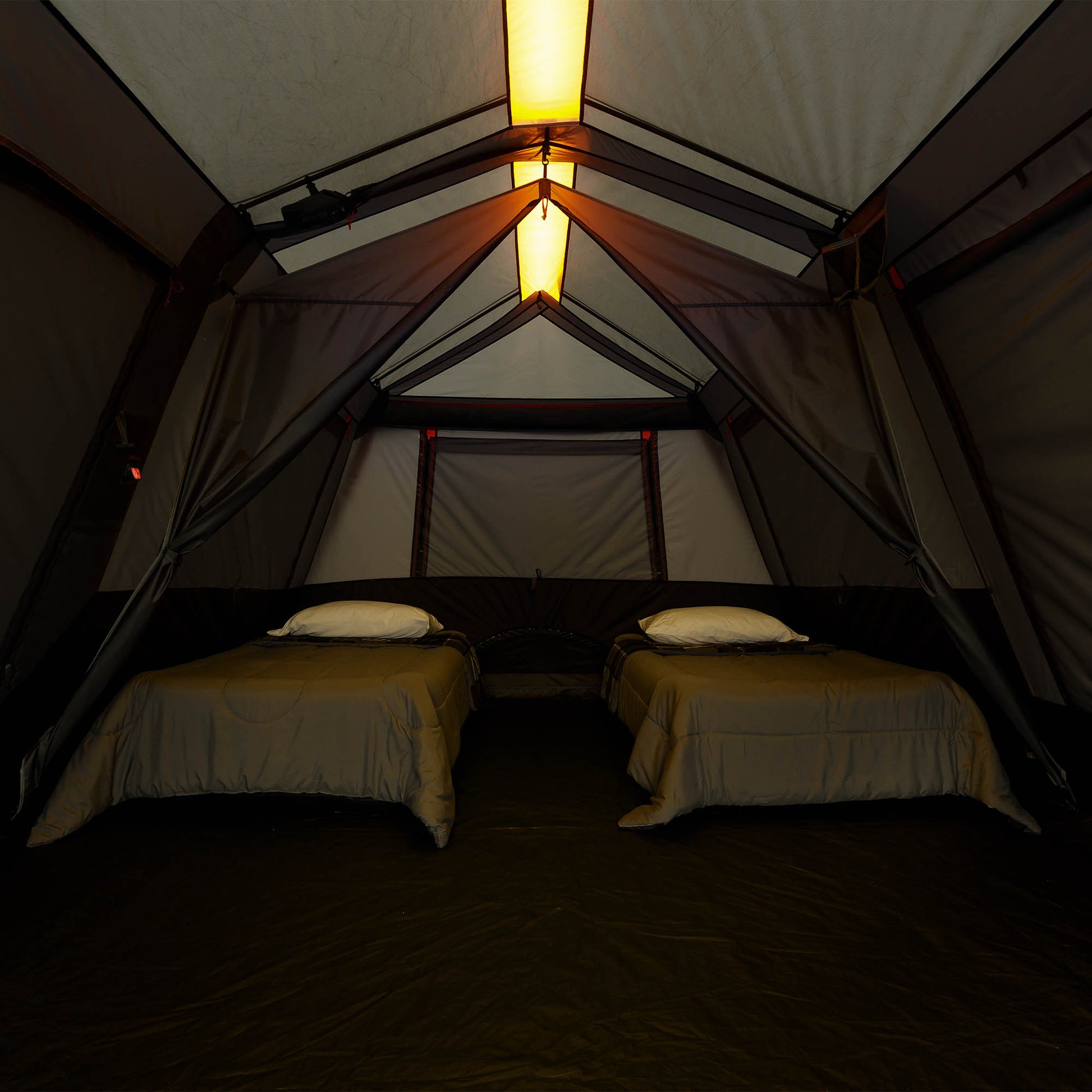 Inside a large camping tent with two single beds, beige bedding, and white pillows, illuminated by warm hanging pendant lights.