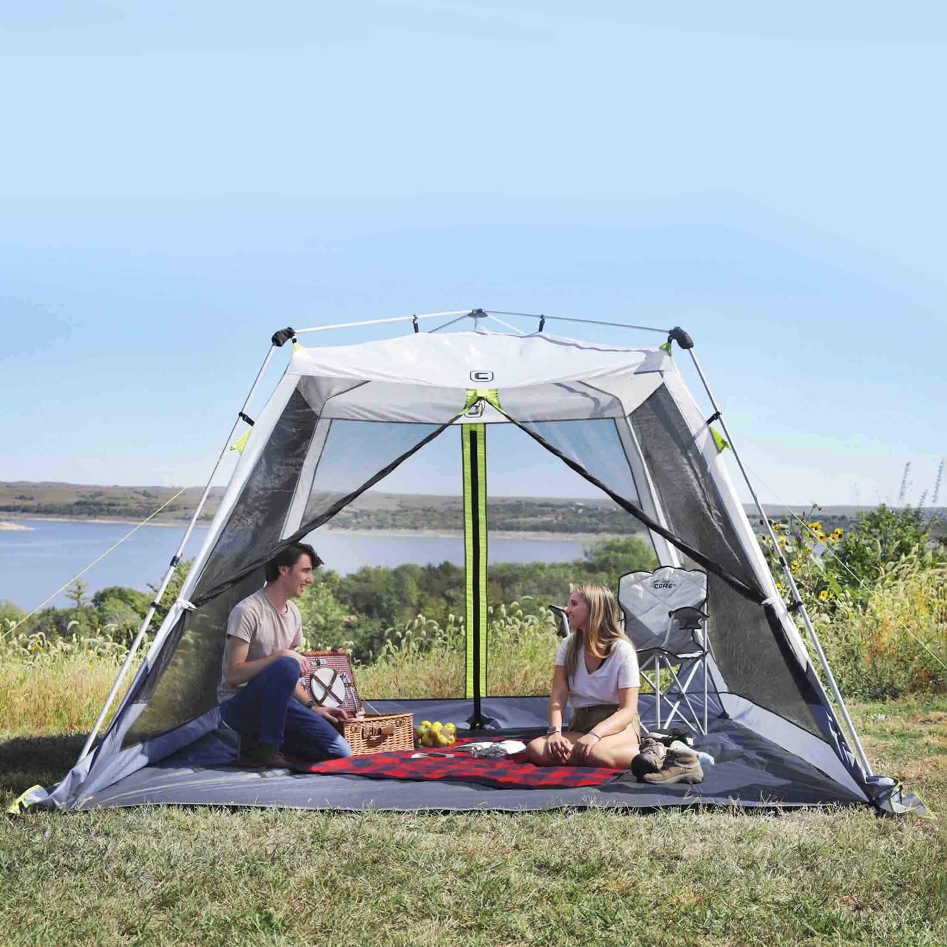 Campers relaxing inside CORE 10x10 instant screen house with mesh walls, enjoying shade and bug-free comfort by the lake.