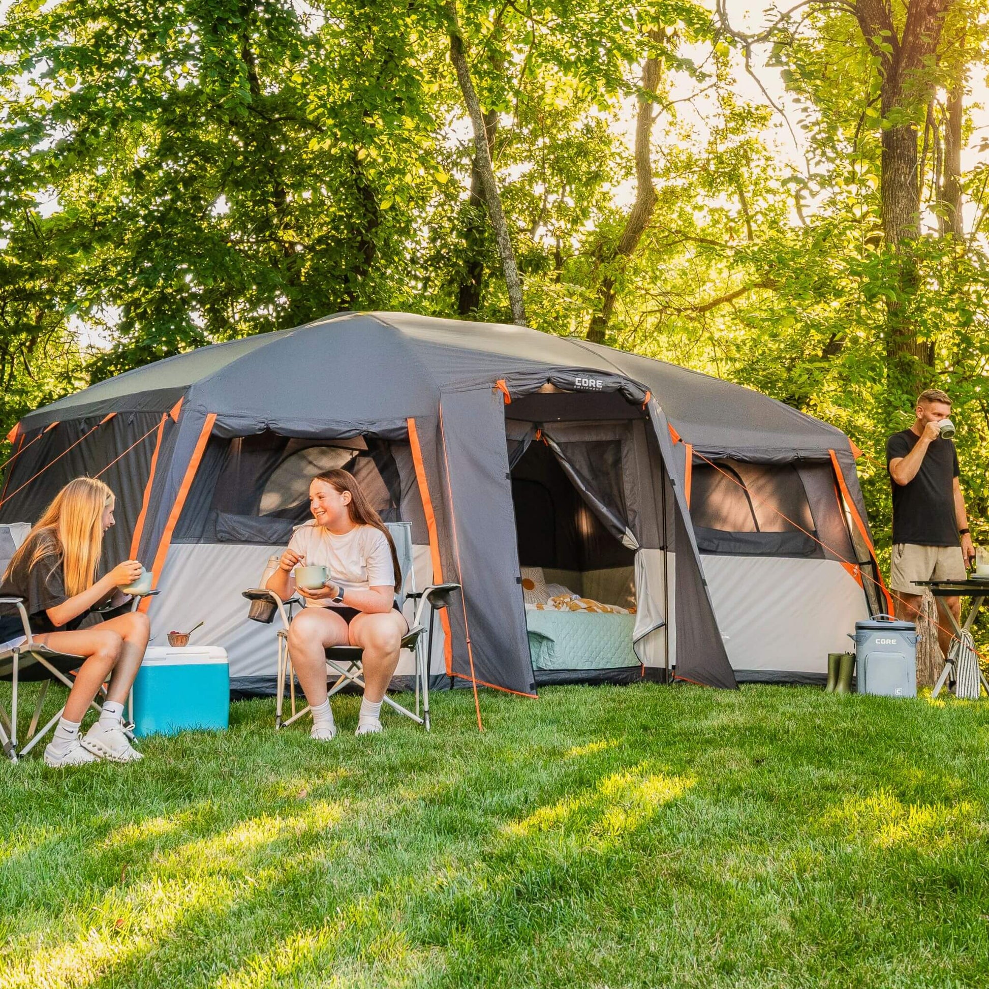 CORE 12 person straight wall cabin tent with full rainfly set up at a campsite with campers sitting outside and gear nearby on grass.