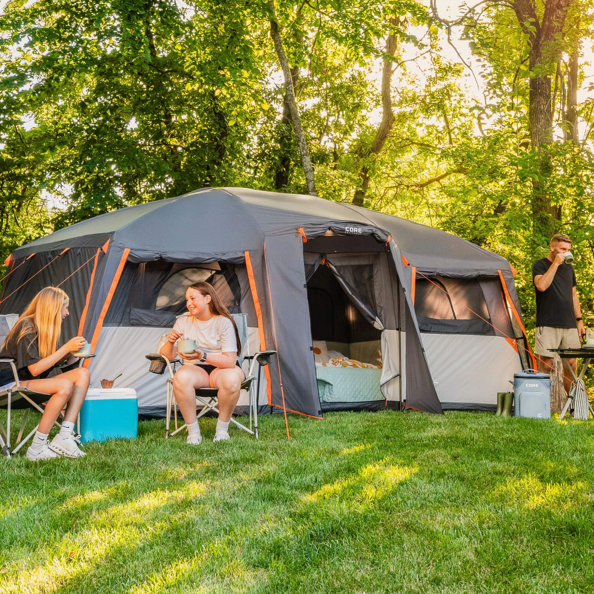 CORE 12 person straight wall cabin tent with full rainfly set up at a campsite with campers sitting outside and gear nearby on grass.