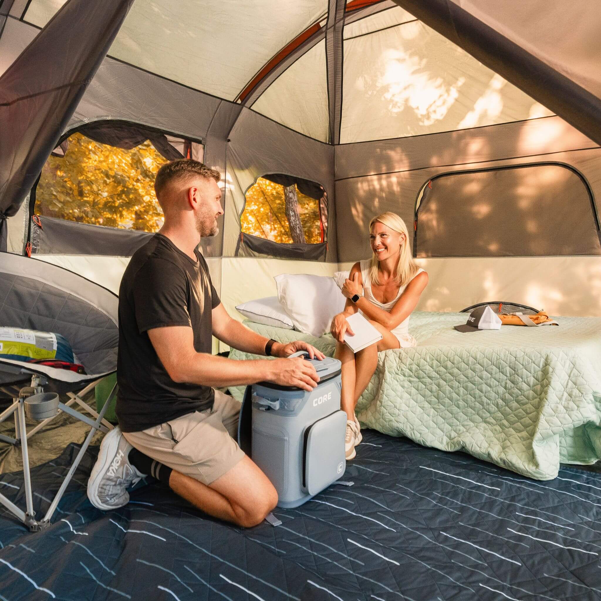 Campers relaxing inside a CORE 12 person straight wall cabin tent with bedding and a CORE soft cooler, enjoying a comfortable campsite interior together.