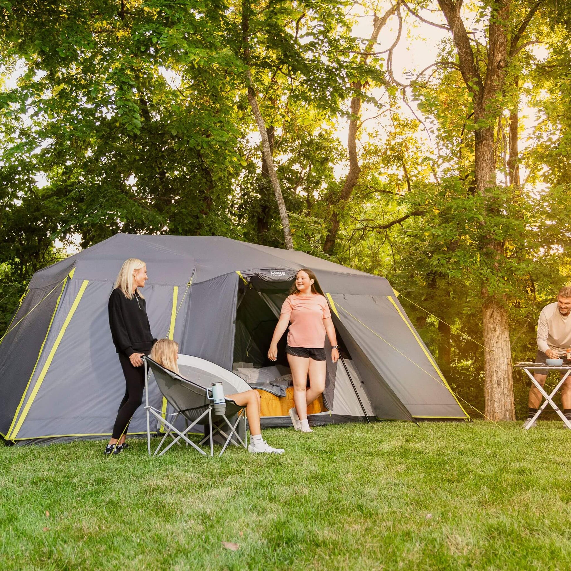 Campers relaxing outside a CORE 12 person instant cabin tent with full rainfly set up on grass with chairs and bedding, enjoying time together at a wooded campsite.