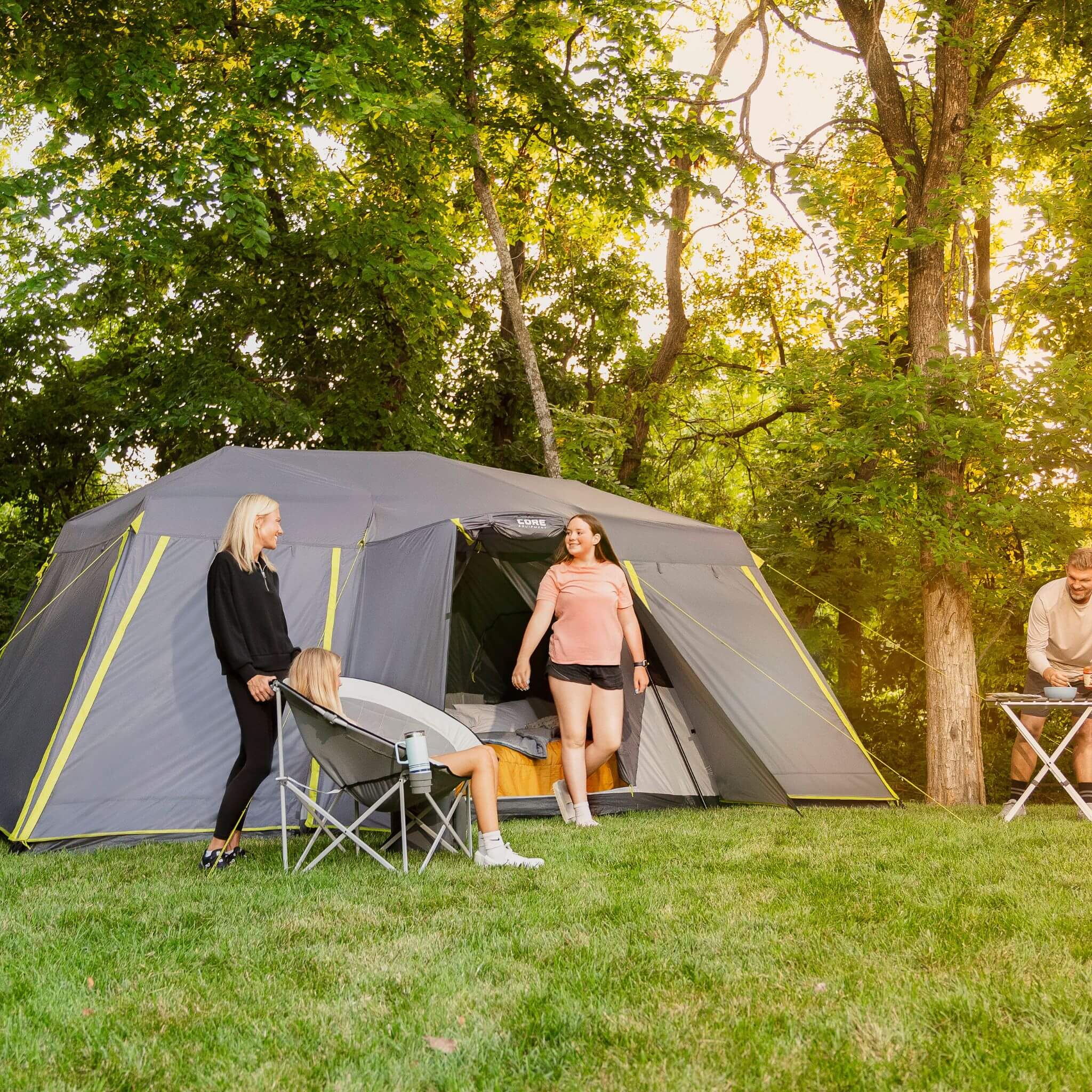 Campers relaxing outside a CORE 12 person instant cabin tent with full rainfly set up on grass with chairs and bedding, enjoying time together at a wooded campsite.