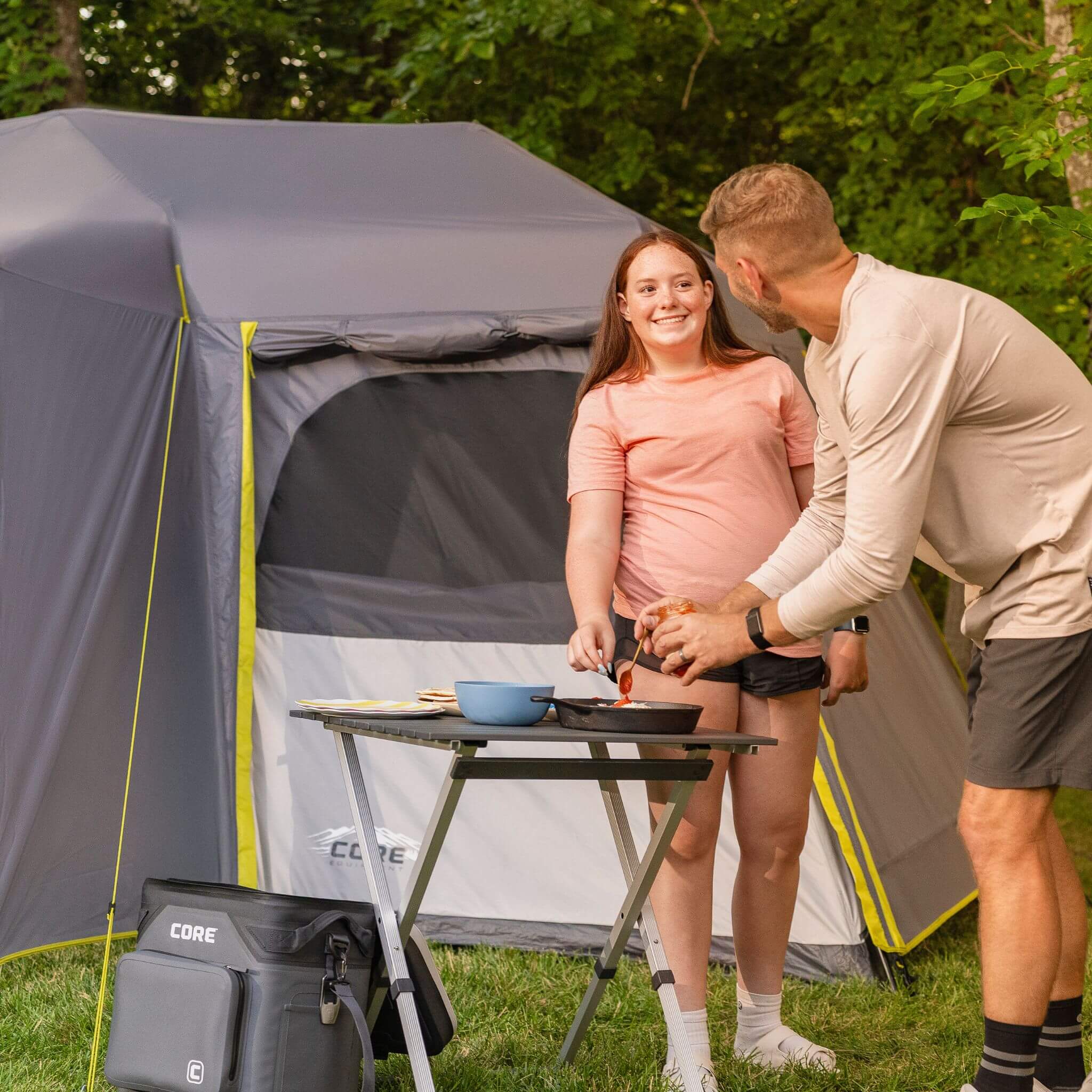 Father and daughter cooking at a campsite beside a CORE 12 person instant cabin tent with full rainfly and a CORE soft cooler and camp table set up on grass.