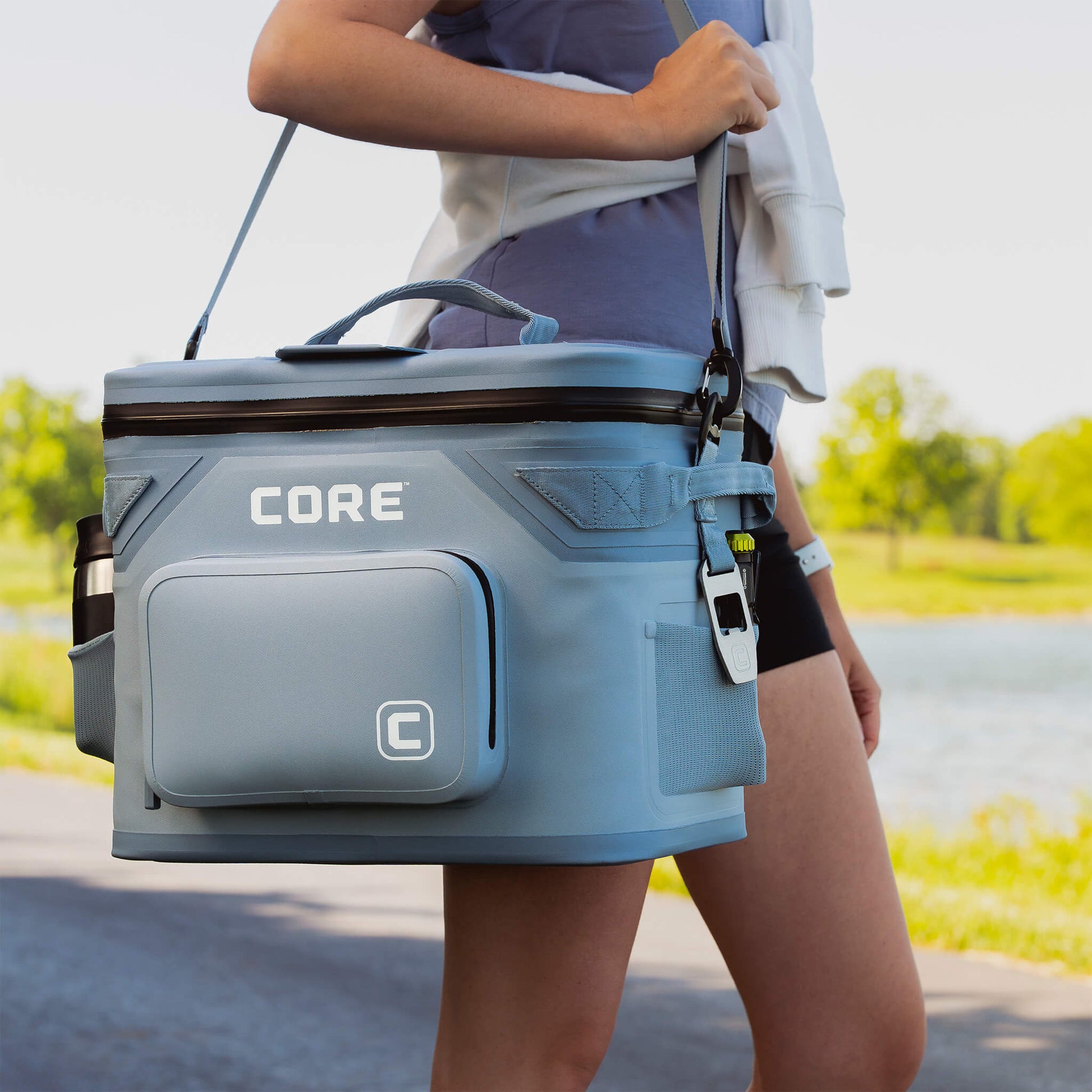 Person carrying CORE Climakeep 18 can steel blue cooler with shoulder strap and side bottle holder near water.