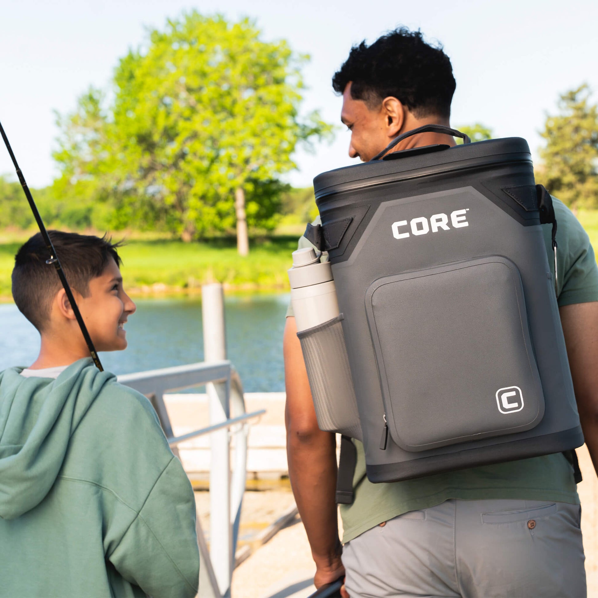 Man carrying CORE Climakeep 24 can charcoal backpack cooler with shoulder strap and side bottle holder while fishing with child.