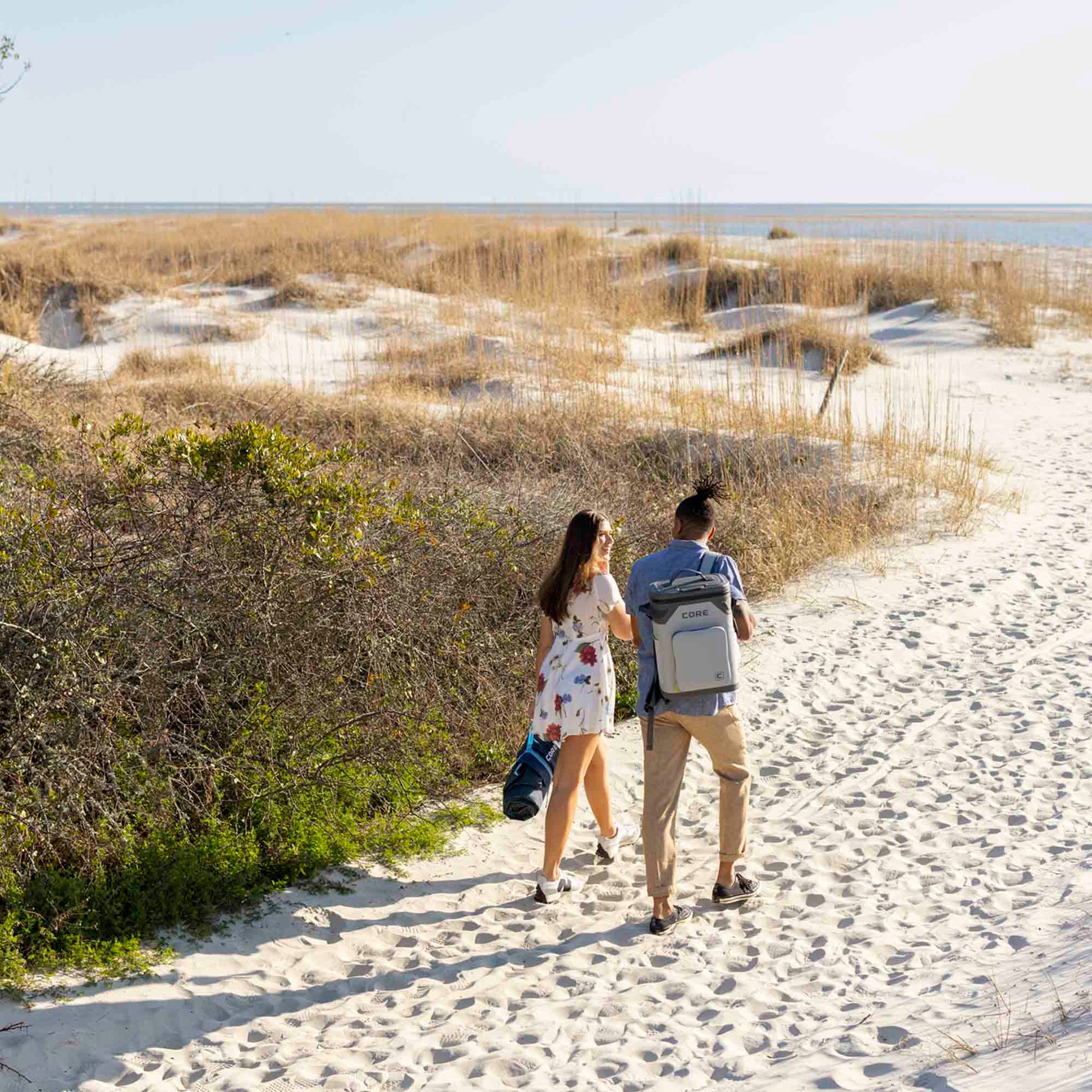 Couple walking on sandy beach, man carrying CORE Climakeep 24 can backpack cooler in shadow gray.