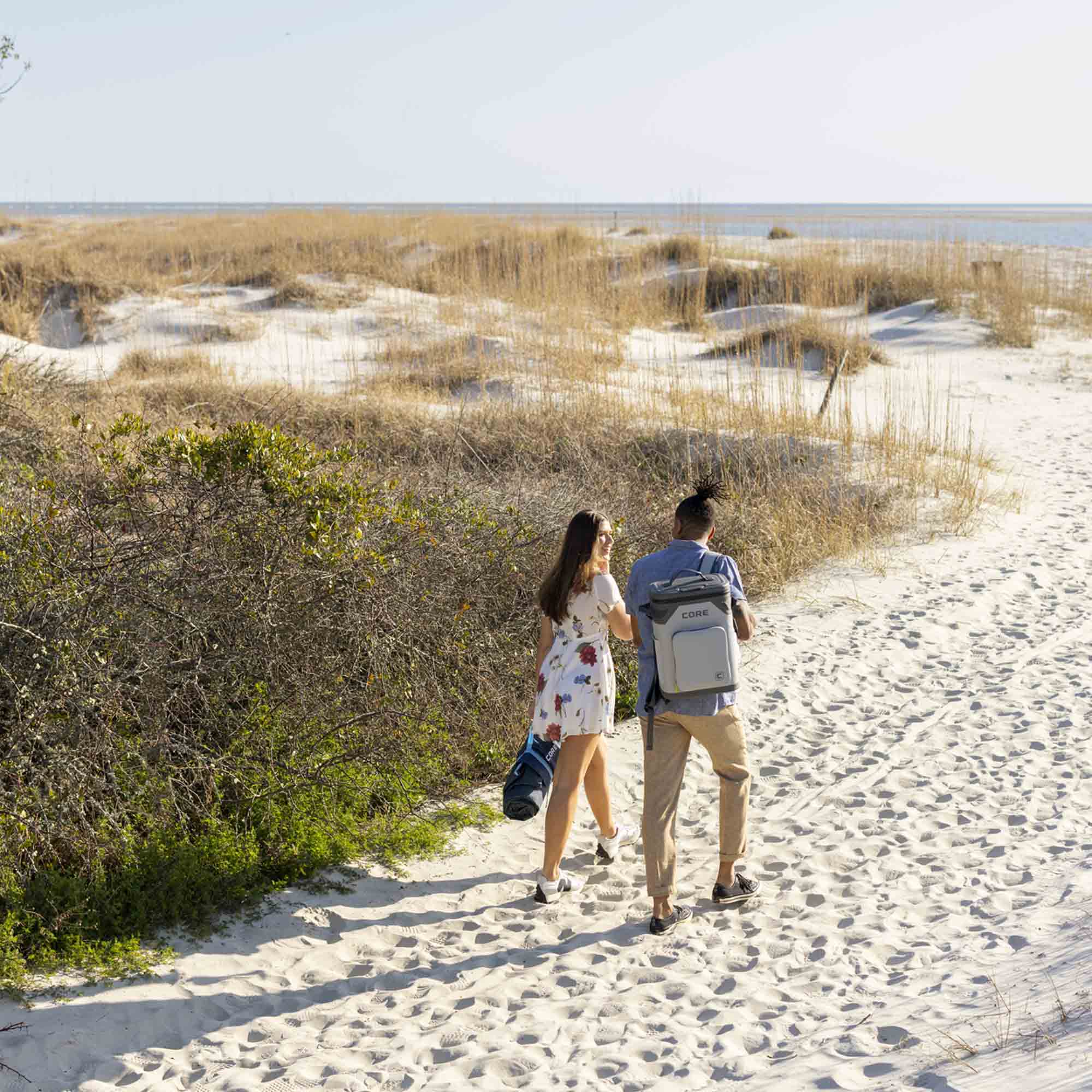 Couple walking on sandy beach, man carrying CORE Climakeep 24 can backpack cooler in shadow gray.