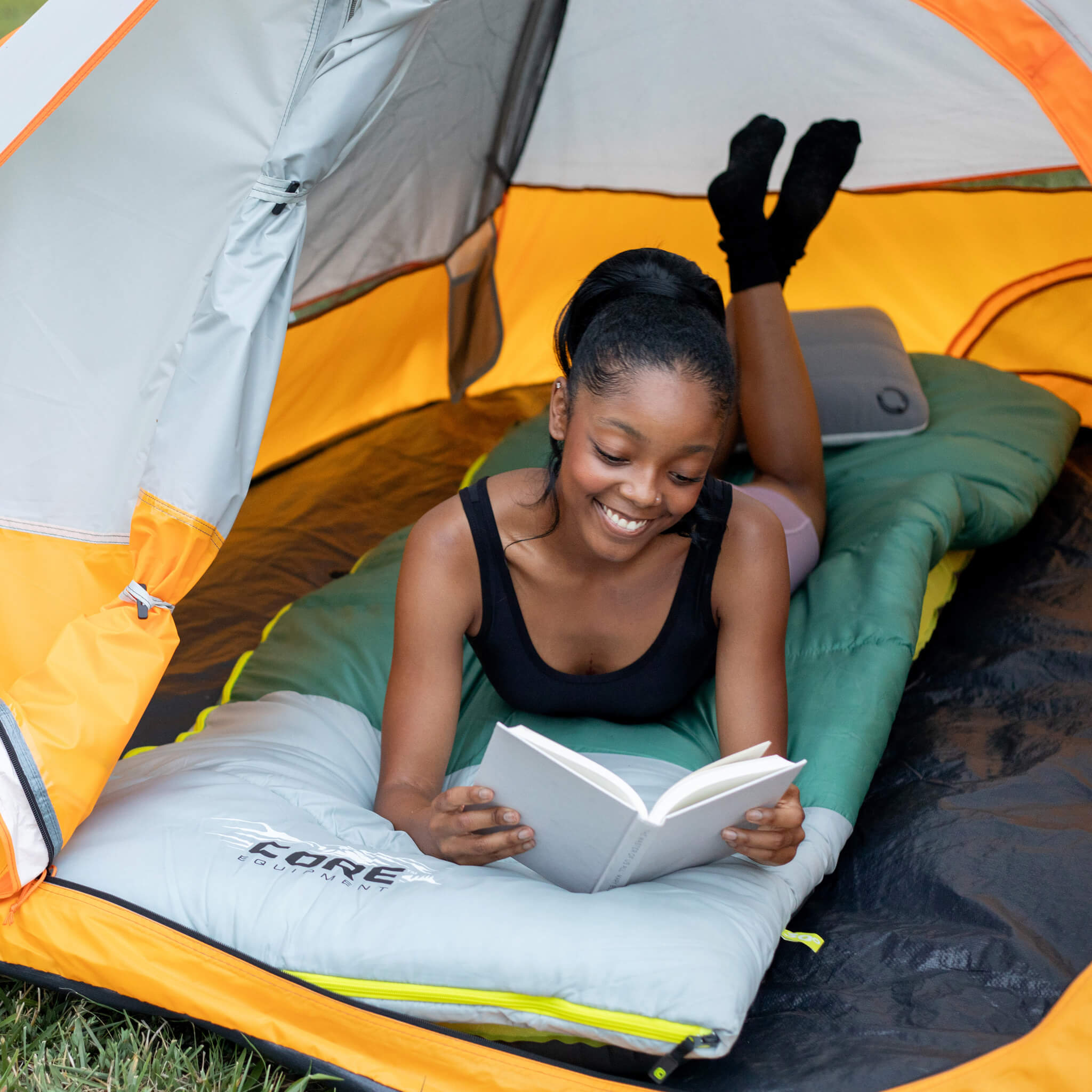 A camper reading inside a CORE 3 person dome tent while lying on a CORE 30 degree sleeping bag.