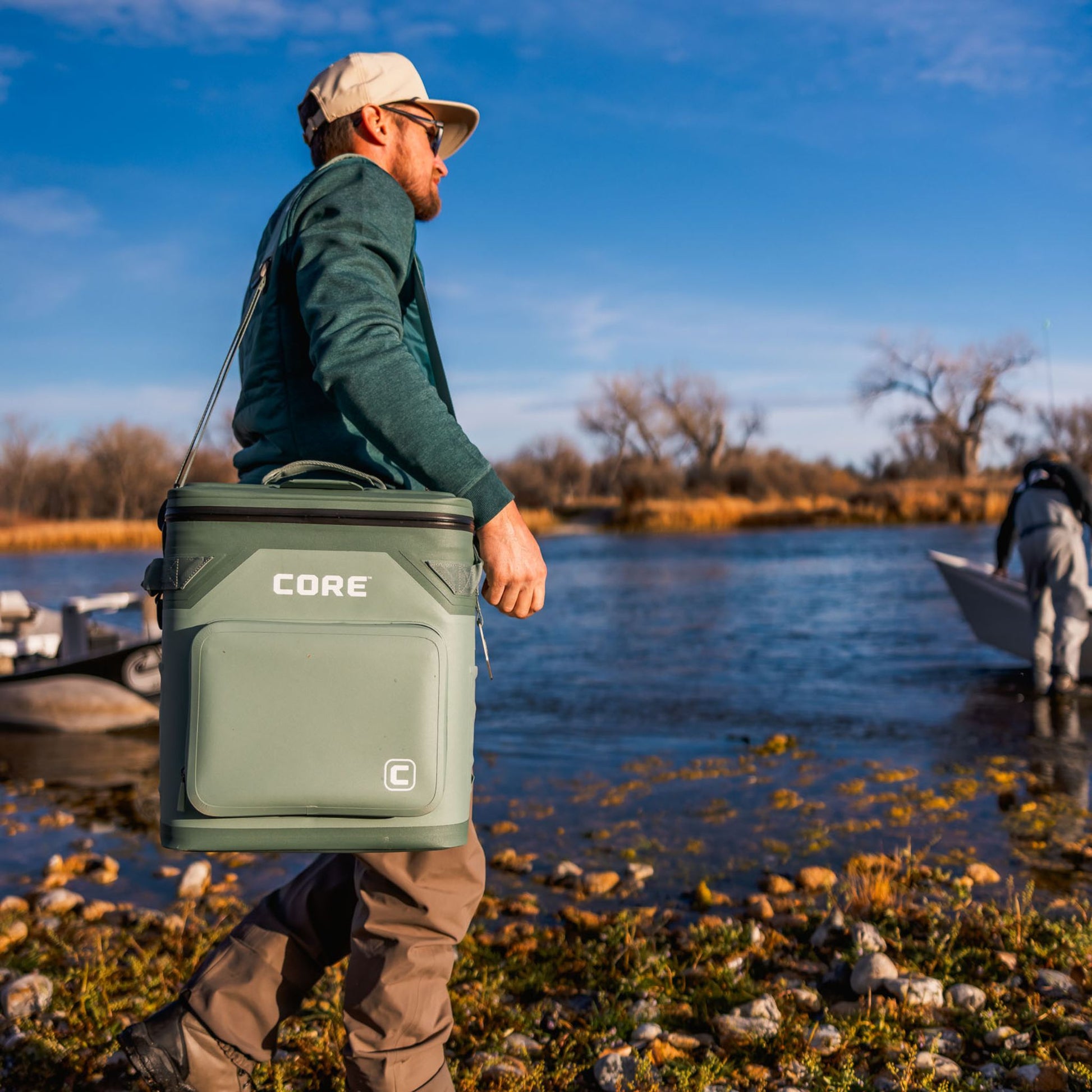 A man is holding a green cooler labeled 'CORE' by a lake with another person in the background.