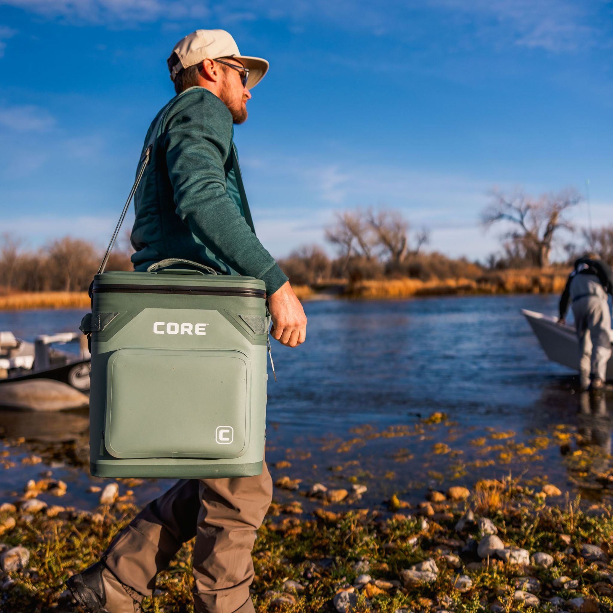 A man is holding a green cooler labeled 'CORE' by a lake with another person in the background.