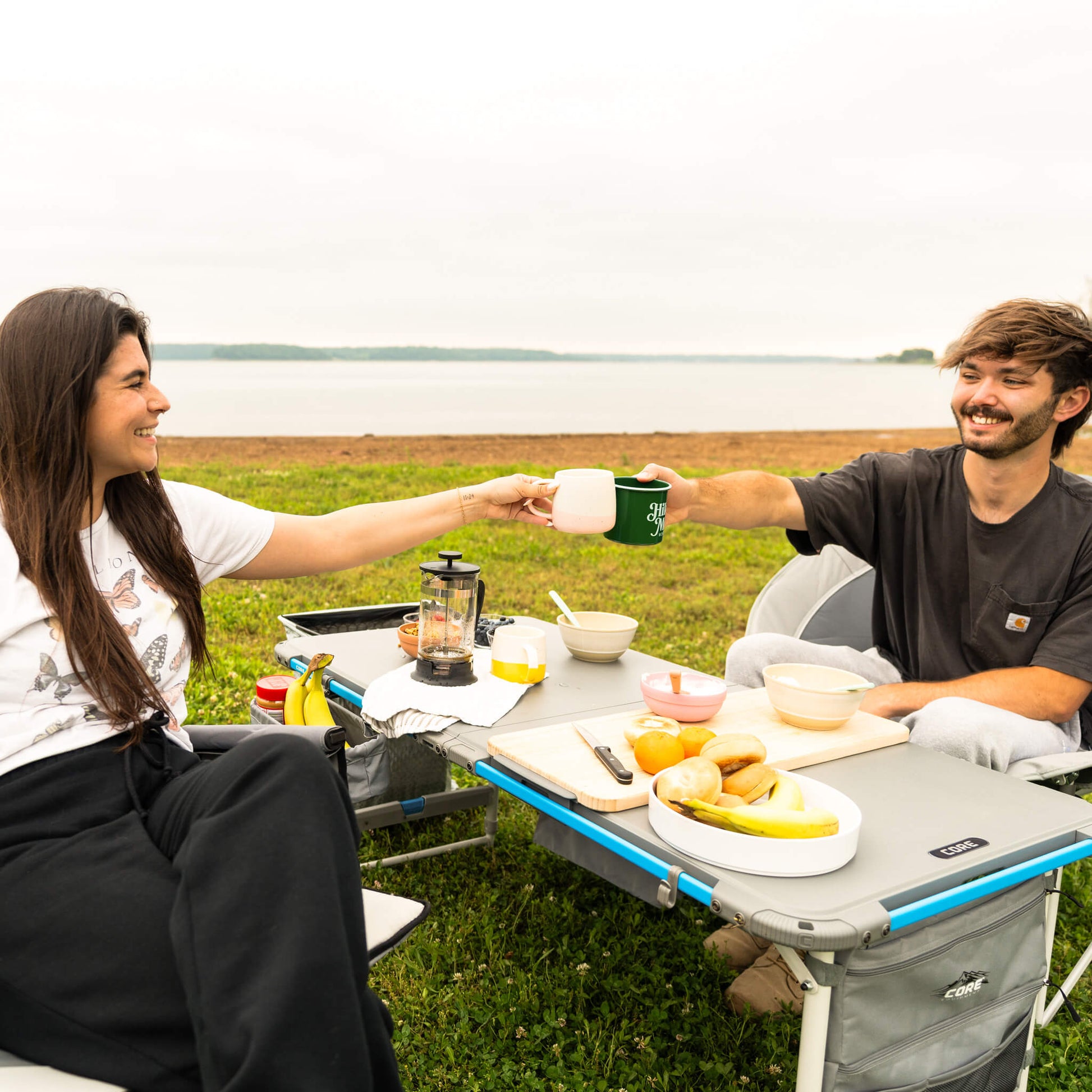 Two campers are enjoying a meal outdoors using the CORE folding camp table with accessories and storage.