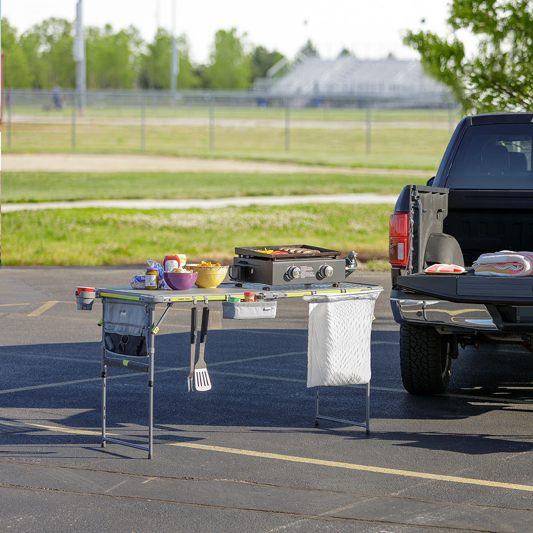 Portable grill setup with food items on a table next to a pickup truck in an outdoor setting.